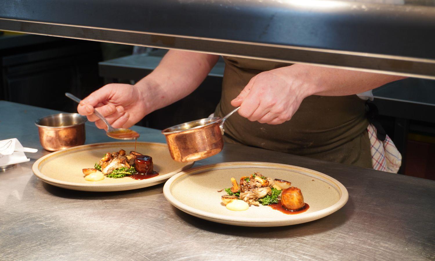 Chef's hands pouring sauce over beautifully prepared dishes on a kitchen pass