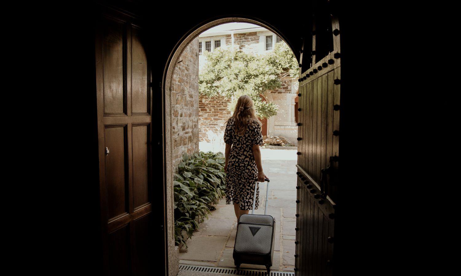 Woman rolling suitcase out of hotel door into sunny courtyard