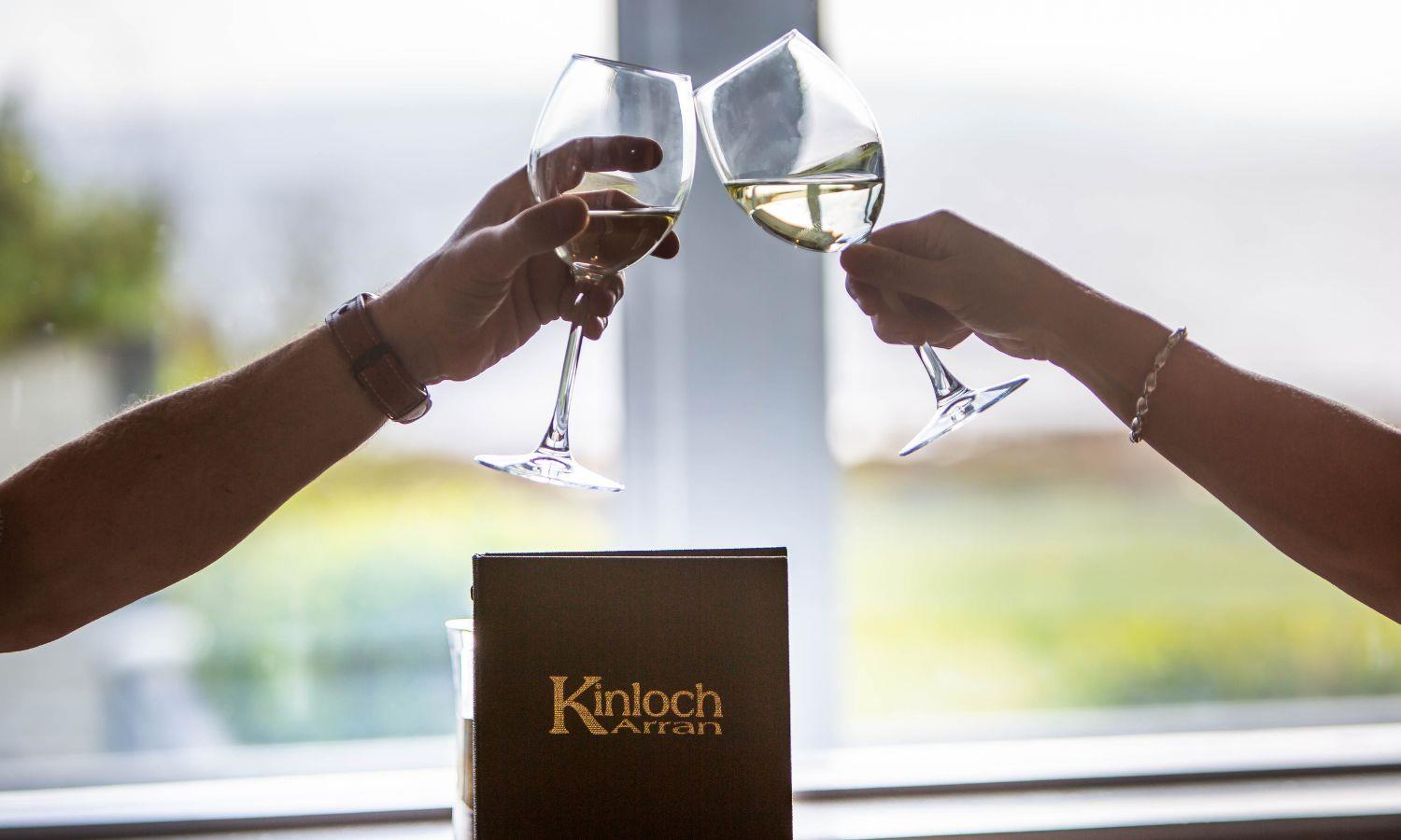 Two people doing a 'cheers' with glasses of white wine with Kinloch Hotel menu in background
