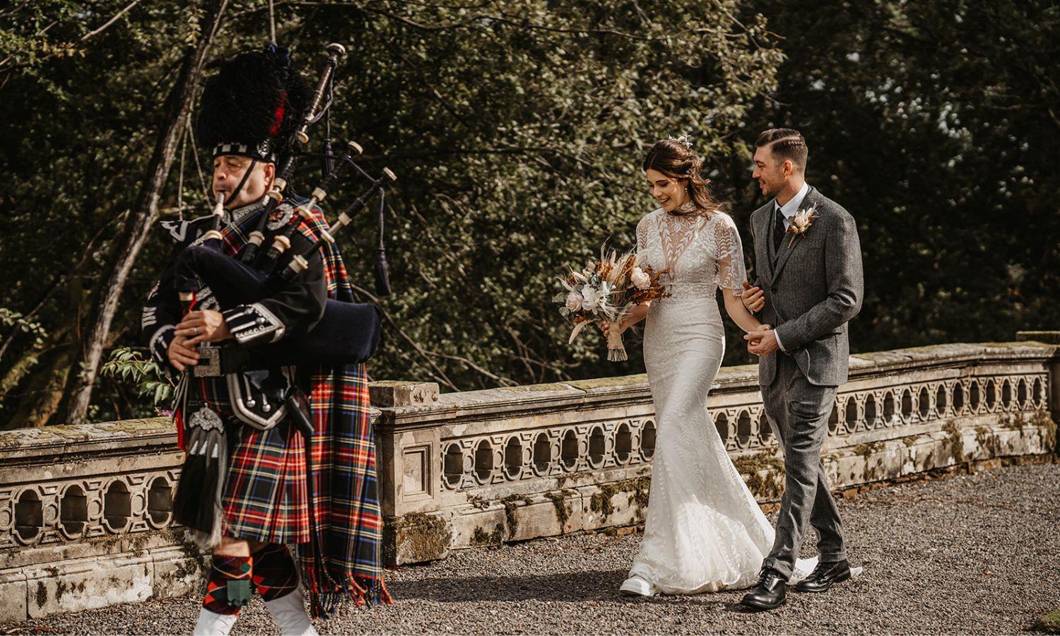 Bride and groom walking behind a bagpiper through the castle ground
