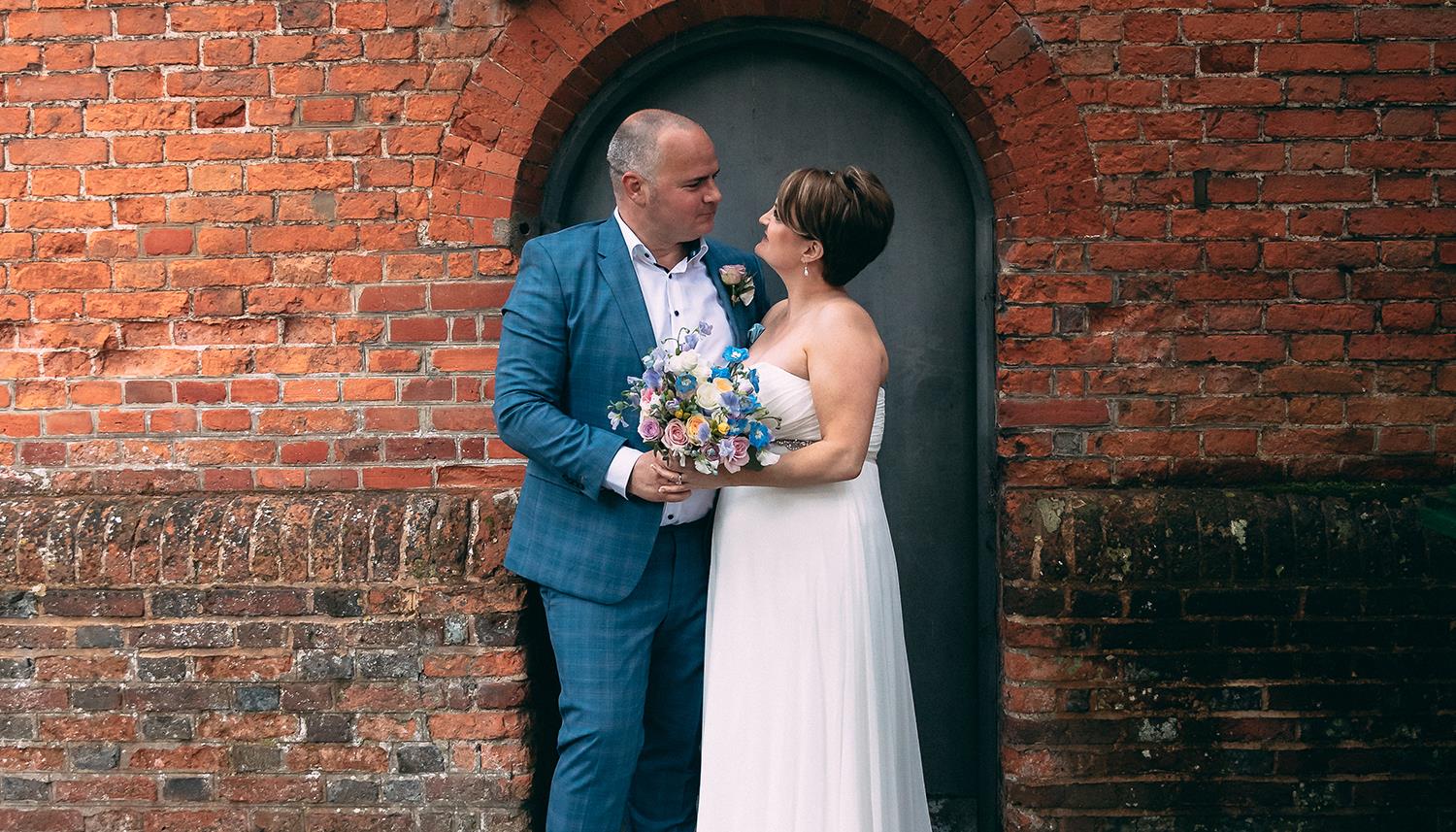 Couple with bouquet standing in front of arched door in brick wall. Photo Credit: F J West 