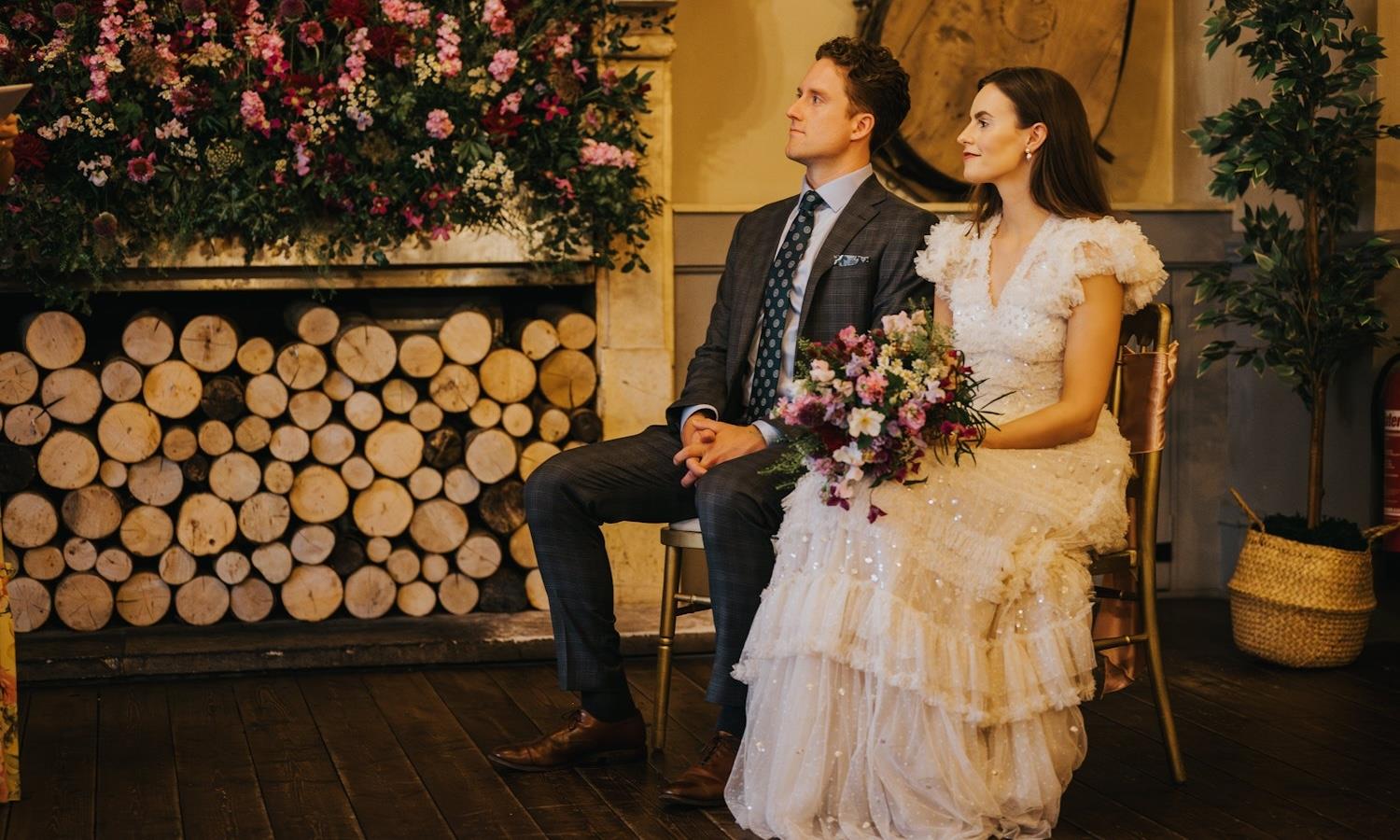 Bride & Groom sat infront of a fire place filled with red and pink flowers.
