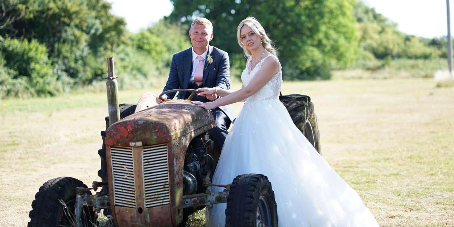 Groom sat on a vintage tractor in field with bride stood next to him (c) Jamie McDine