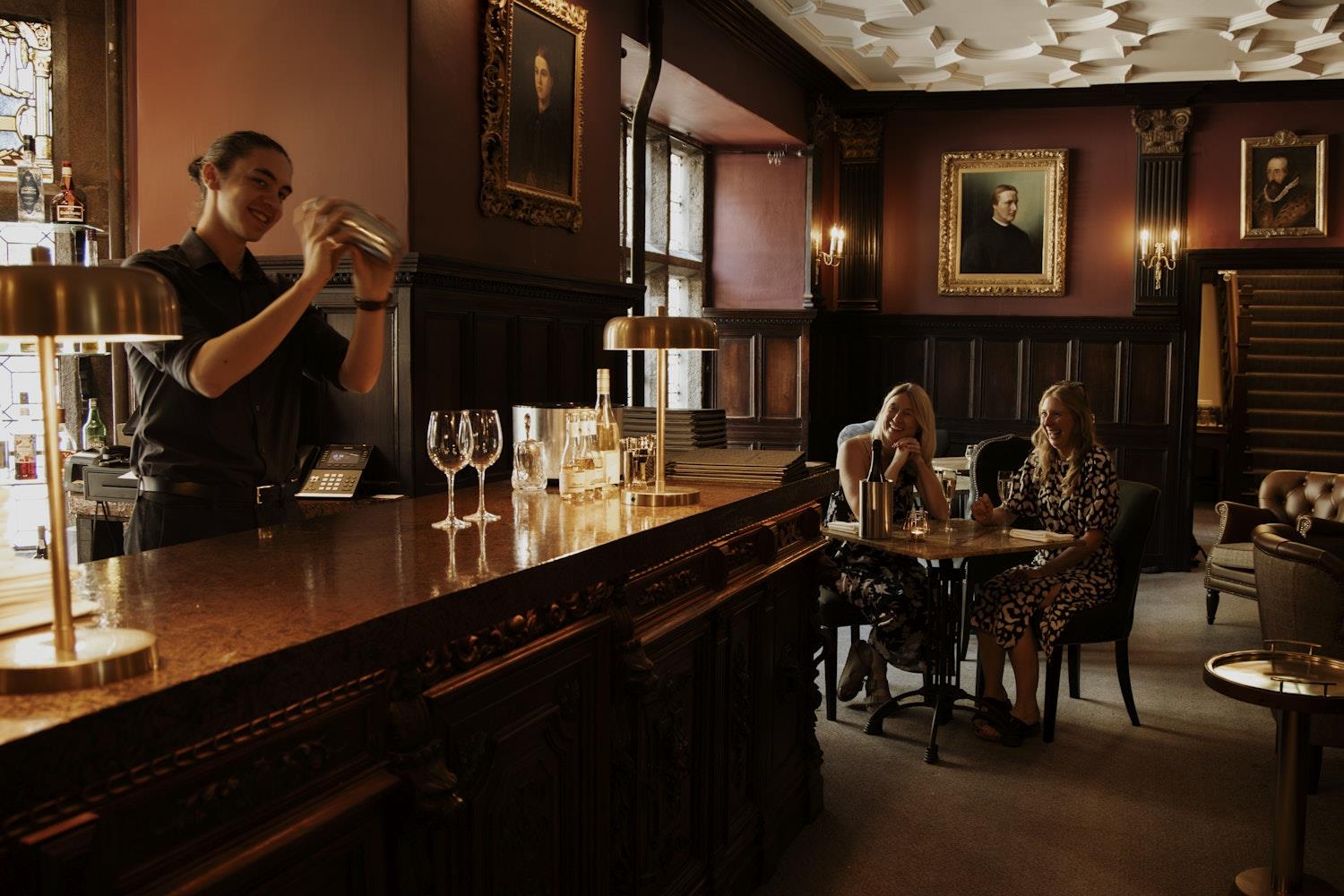 Bartender makes two cocktails as two guests look on 