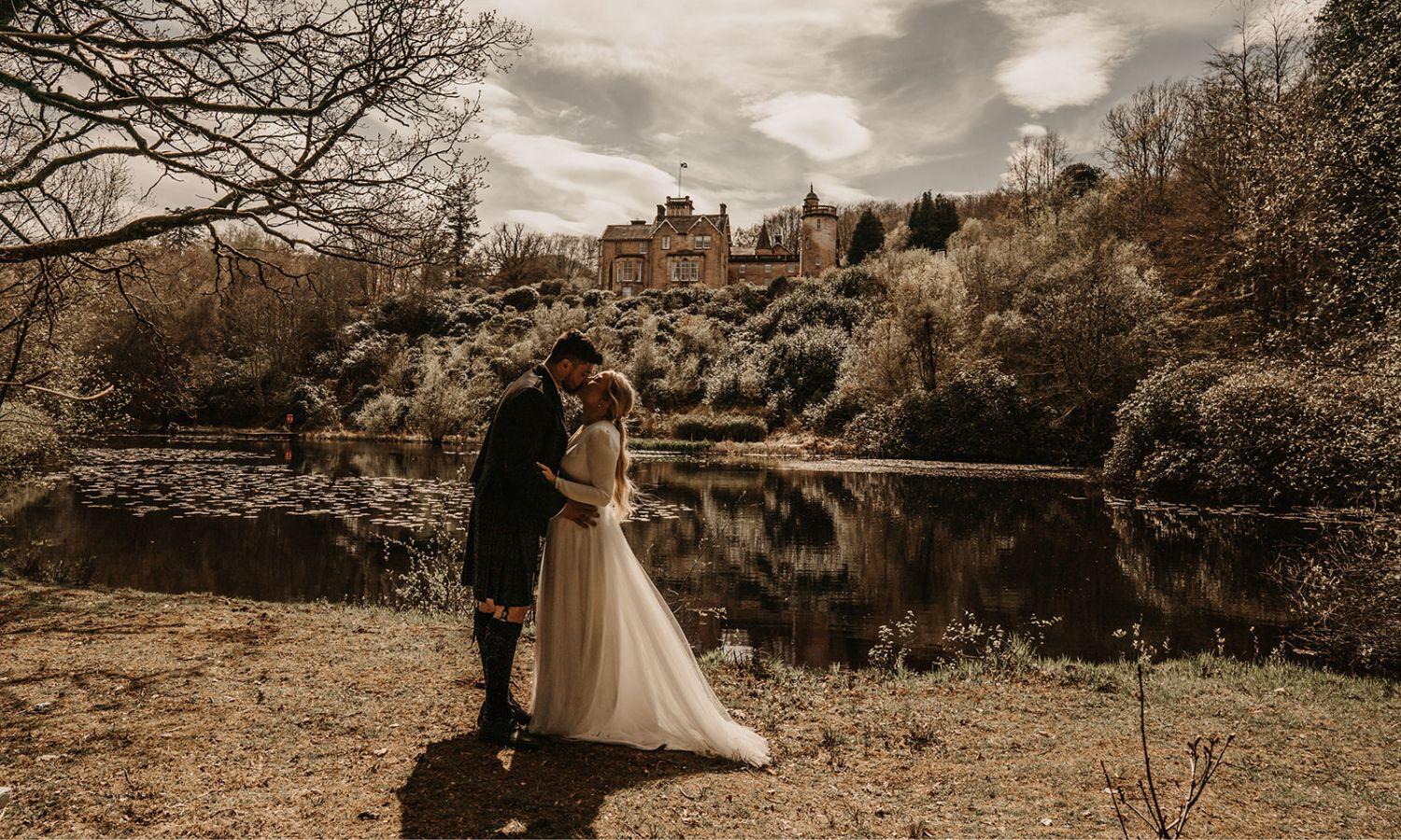 Bride and groom embracing beside the private lake at Auchen Castle, surrounded by woodland