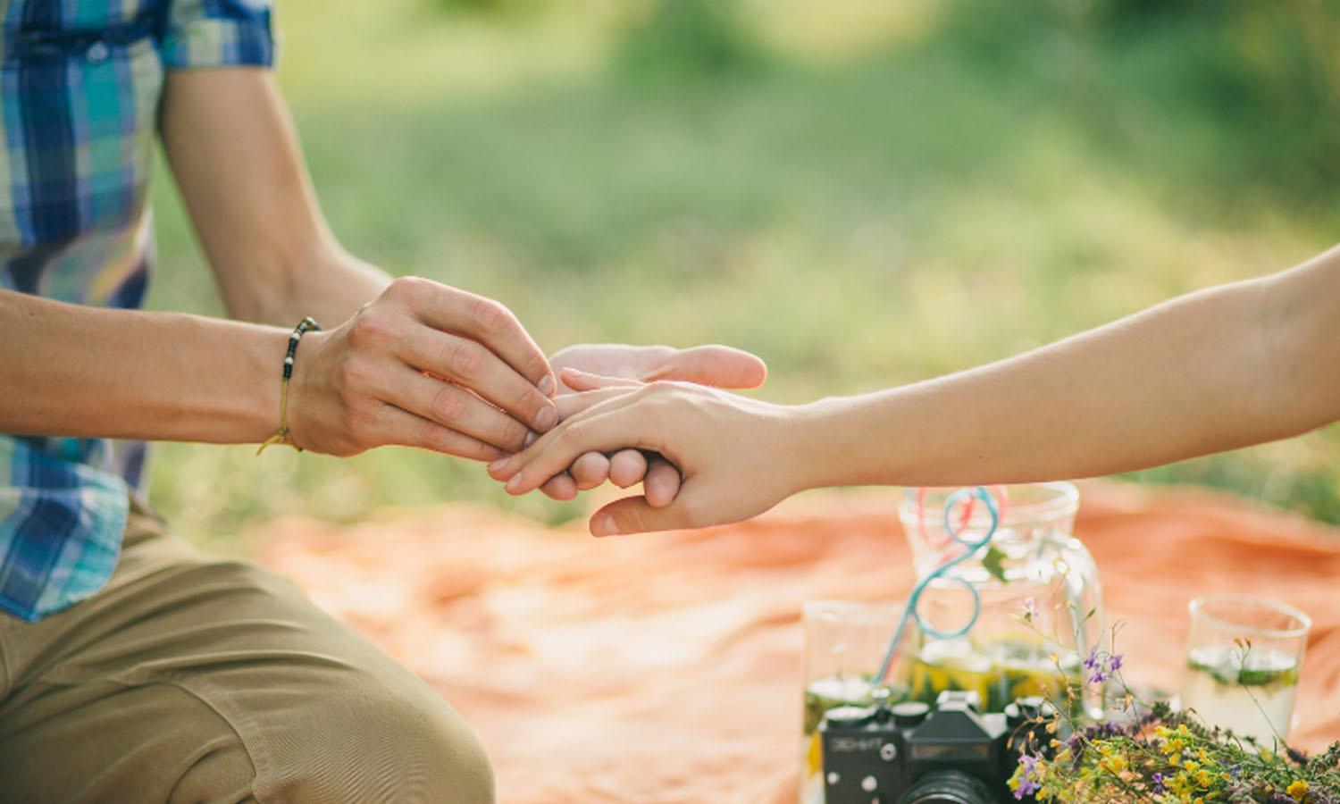 Man and putting ring on women's finger sitting on picnic blanket