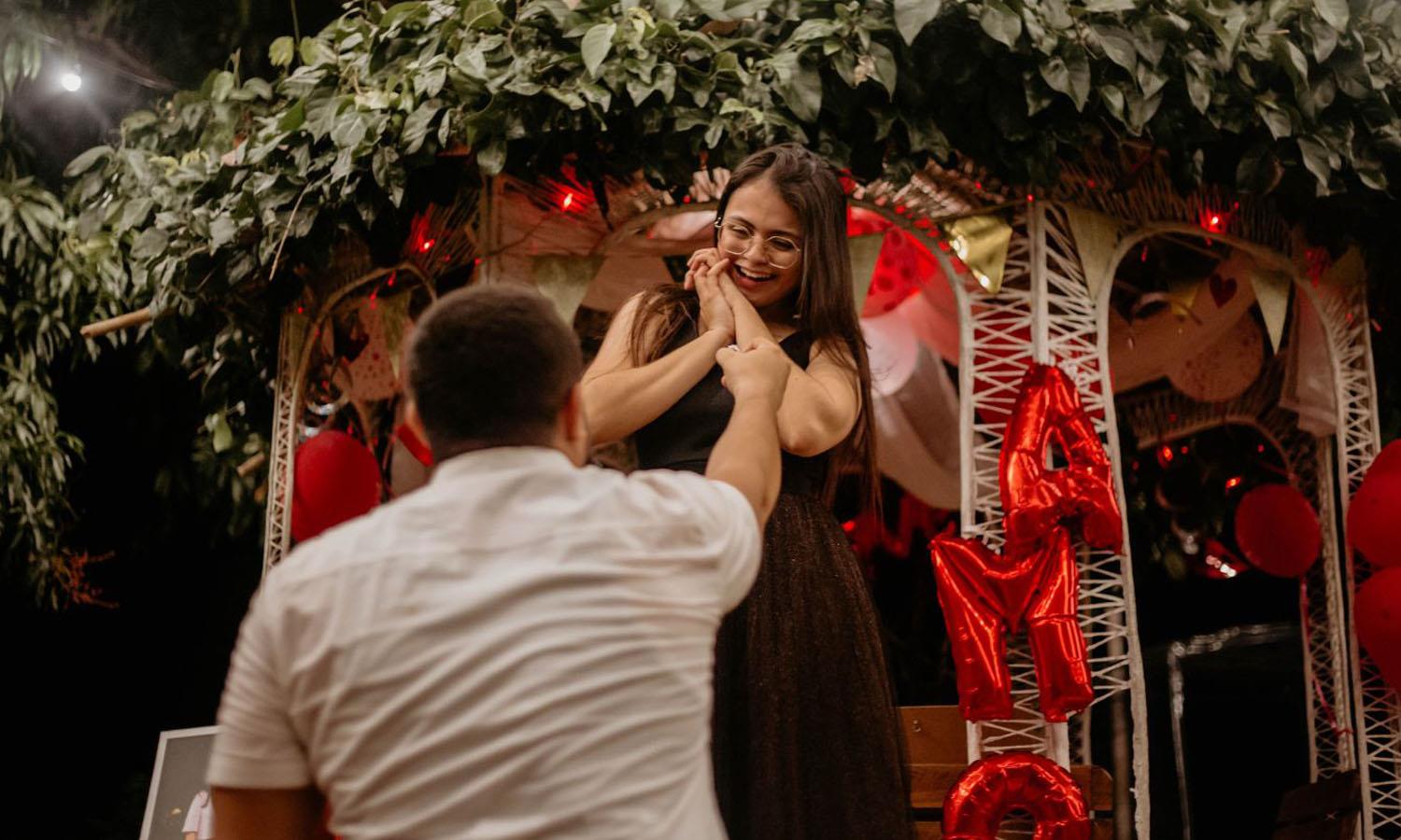 Man proposing to woman in front of a white arbor with red balloons